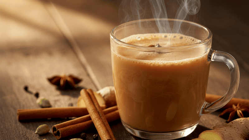 A steaming glass cup of masala chai surrounded by whole spices on a rustic wooden surface