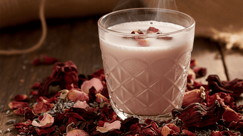 Dried rose petals, lavender buds, and hibiscus flowers scattered around a glass cup of pink-hued floral chai