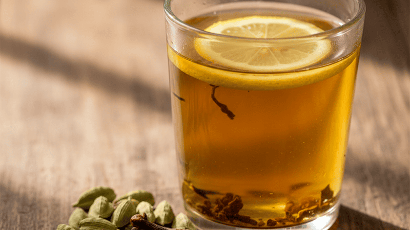 A clear glass of golden sulaimani tea with a lemon wedge and whole cardamom pods on a dark wooden table