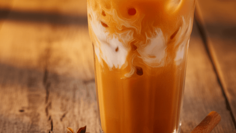 A glass of bright orange Thai iced tea next to a cup of masala chai with star anise and cardamom scattered between them